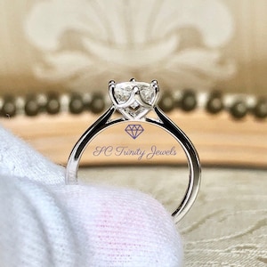 May include: A silver-toned ring featuring a single clear, faceted stone held in a six-prong setting. The band is smooth and polished. The text SC Trinity Jewels and a small diamond icon are printed in blue on the center of the ring band.