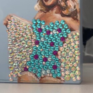 May include: A light blue case featuring a painted portrait of a woman with blonde hair. The case is embellished with iridescent and turquoise rhinestones, arranged in a pattern. The design includes magenta accents.