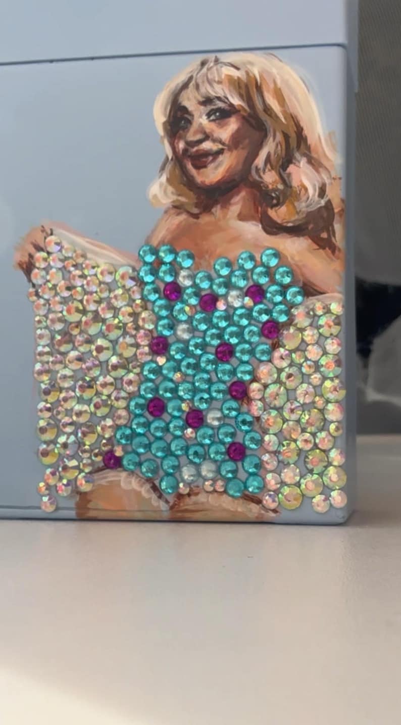 May include: A light blue case featuring a painted portrait of a woman with blonde hair. The case is embellished with iridescent and turquoise rhinestones, arranged in a pattern. The design includes magenta accents.