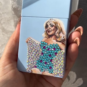 May include: A light blue cigarette case featuring a painted portrait of a woman with blonde hair. The woman is wearing a turquoise dress adorned with sparkling rhinestones and magenta accents.