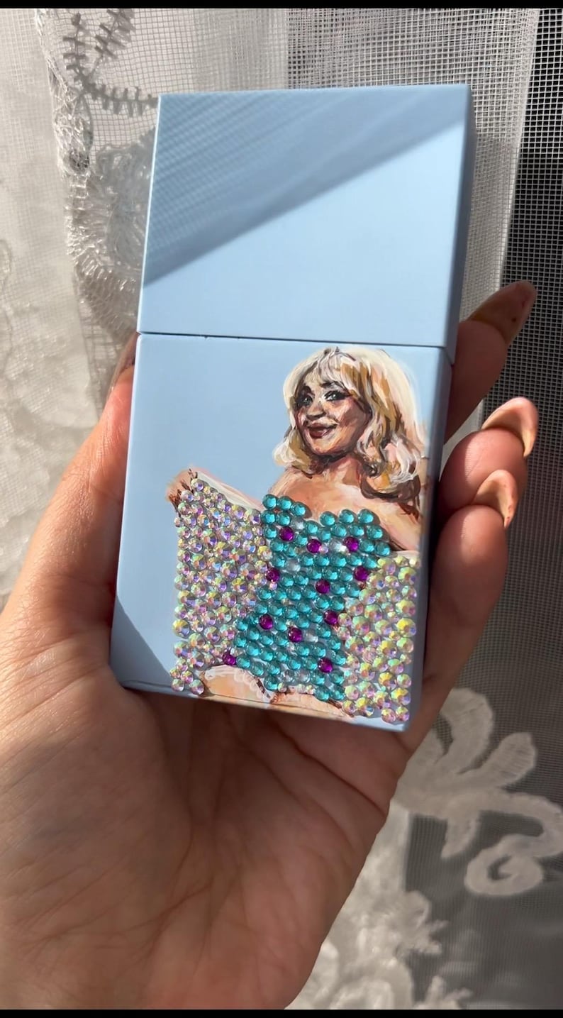 May include: A light blue cigarette case featuring a painted portrait of a woman with blonde hair. The woman is wearing a turquoise dress adorned with sparkling rhinestones and magenta accents.