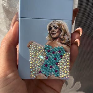 May include: A light blue cigarette case featuring a painted portrait of a woman with blonde hair. The woman is wearing a turquoise dress adorned with sparkling rhinestones and purple accents. The case is held in a hand.