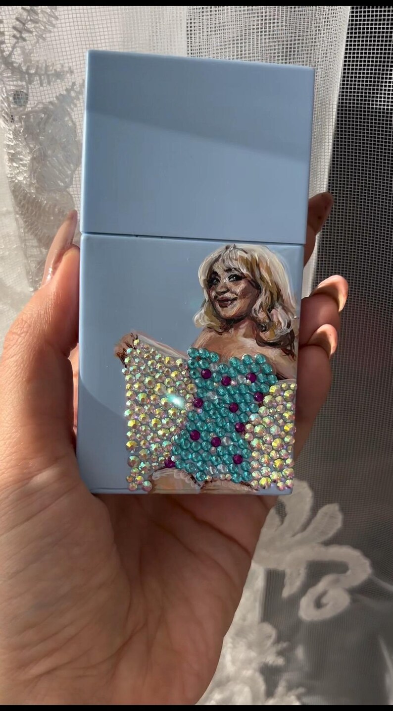 May include: A light blue cigarette case featuring a painted portrait of a woman with blonde hair. The woman is wearing a turquoise dress adorned with sparkling rhinestones and purple accents. The case is held in a hand.