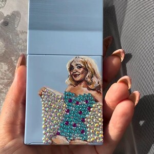 May include: A light blue cigarette case featuring a hand-painted portrait of a woman with blonde hair. The woman is wearing a turquoise dress adorned with sparkling crystals and purple accents. The case is held in a hand.