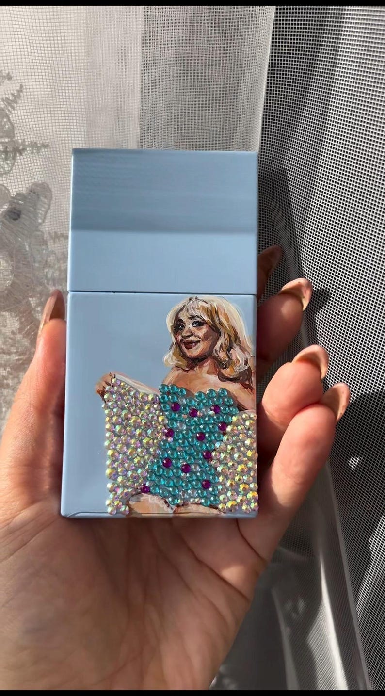 May include: A light blue cigarette case featuring a hand-painted portrait of a woman with blonde hair. The woman is wearing a turquoise dress adorned with sparkling crystals and purple accents. The case is held in a hand.