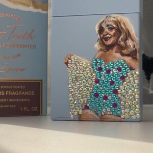 May include: A light blue perfume box with a painted portrait of a woman. The woman is wearing a jeweled top with turquoise and pink accents. The box also has text that reads "Sweet Tooth" and "1 FL. OZ."