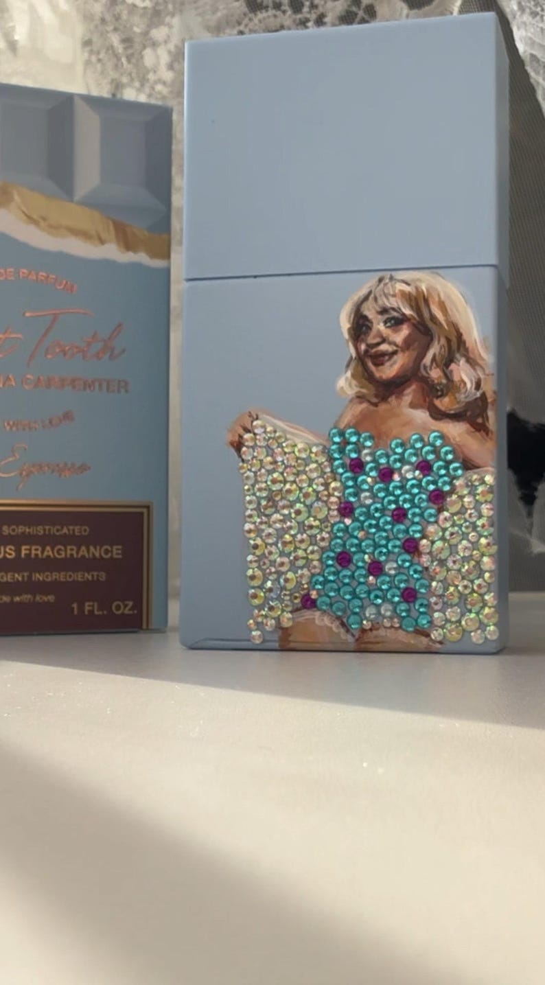 May include: A light blue perfume box with a painted portrait of a woman. The woman is wearing a jeweled top with turquoise and pink accents. The box also has text that reads "Sweet Tooth" and "1 FL. OZ."