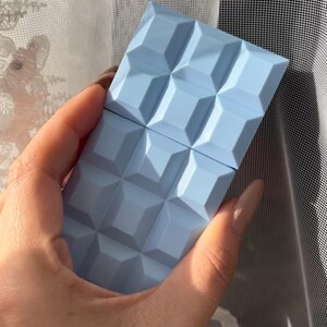 May include: A light blue rectangular object with a geometric pattern, resembling a chocolate bar, is held in a hand. The object's surface is divided into square sections. The background includes a sheer white curtain.