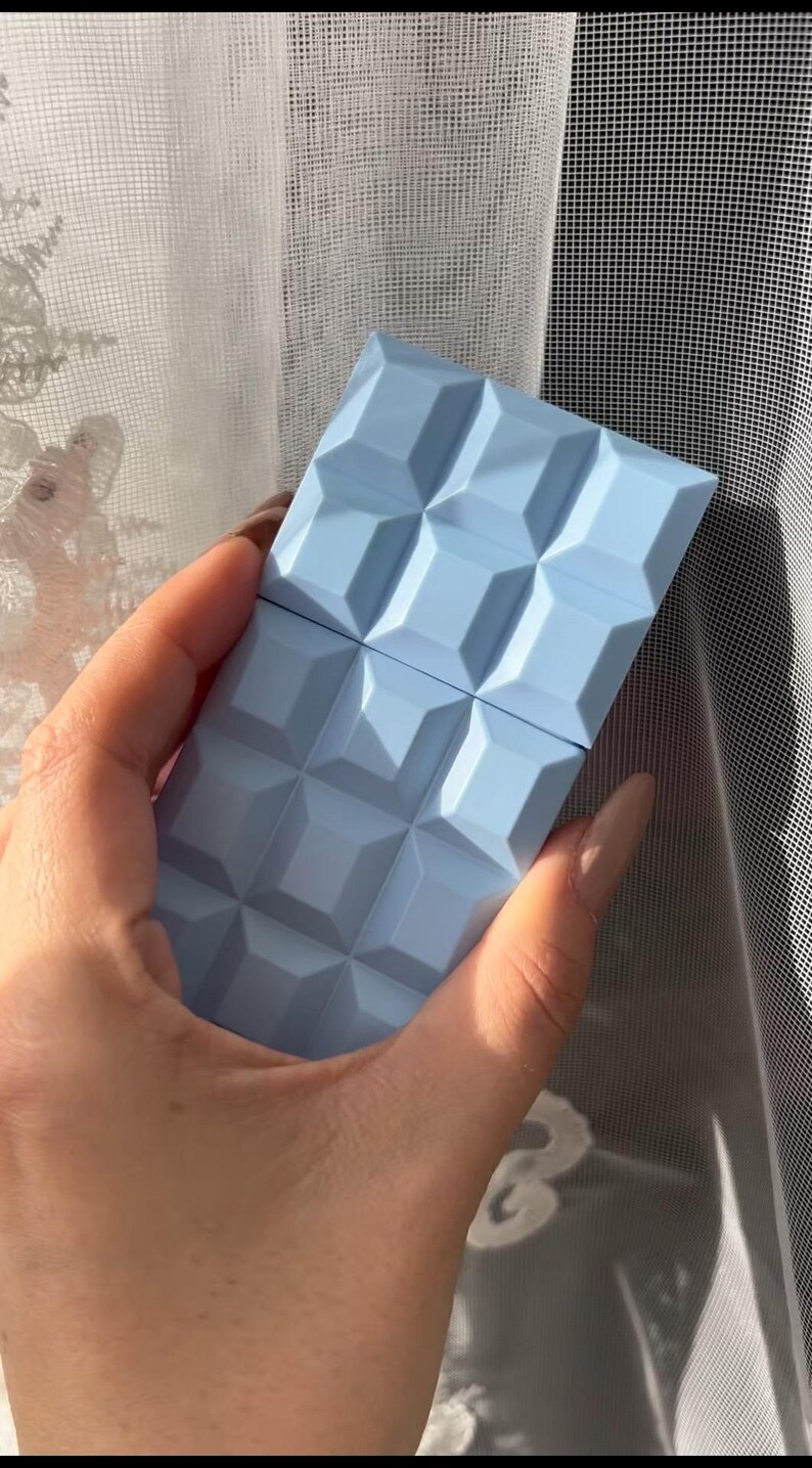 May include: A light blue rectangular object with a geometric pattern, resembling a chocolate bar, is held in a hand. The object's surface is divided into square sections. The background includes a sheer white curtain.