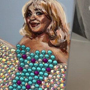 May include: A light blue box featuring a painted portrait of a woman with blonde hair. The woman's dress is adorned with turquoise and purple rhinestones, and the lower portion of the box is covered in iridescent rhinestones.