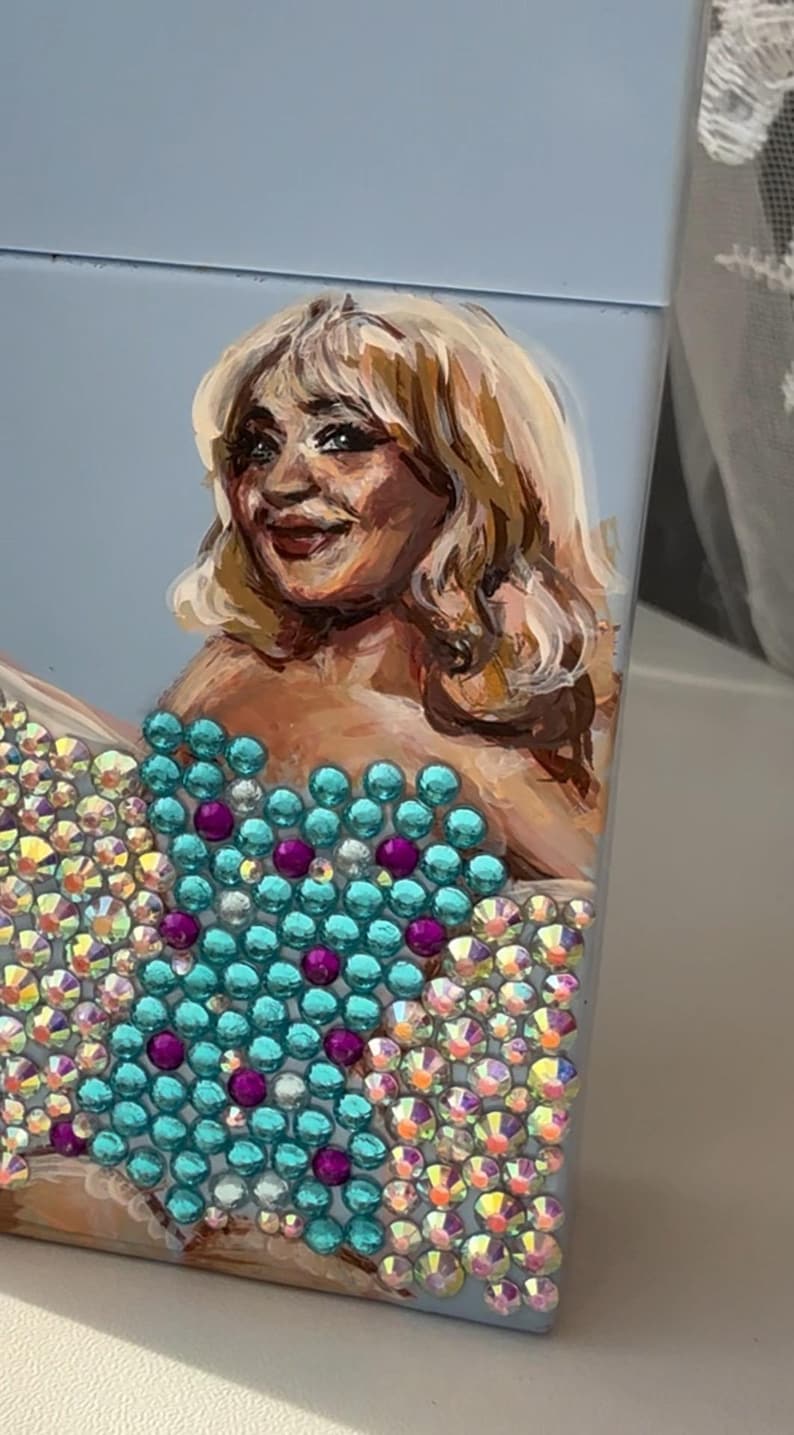 May include: A light blue box featuring a painted portrait of a woman with blonde hair. The woman's dress is adorned with turquoise and purple rhinestones, and the lower portion of the box is covered in iridescent rhinestones.