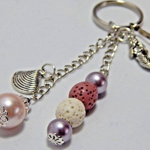 May include: A silver-toned keychain featuring a metal ring, a small seahorse charm, and two dangling chains. One chain holds a silver seashell charm and a pale pink pearl bead. The other chain features a purple pearl, a white lava bead, a dusty rose lava bead, and a purple pearl bead.