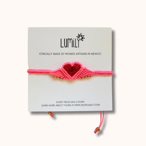 Puede incluir: Una pulsera de macramé rosa con un diseño de corazón y alas, exhibida en una tarjeta blanca. La tarjeta presenta el nombre de la marca "LUMILY" y el texto "ETHICALLY MADE BY WOMEN ARTISANS IN MEXICO." La pulsera tiene detalles dorados y cierre de cuerda.