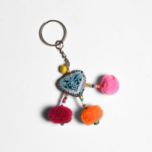 May include: A heart-shaped keychain with a silver ring and chain. The heart is embroidered with a blue design on a black background. It has pink, orange, and red pom-poms and colorful beads. A yellow bead is at the top.