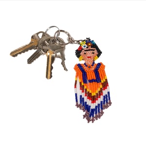 May include: A beaded keychain featuring a Frida Kahlo design, with a colorful poncho in orange, blue, white, and brown. The keychain is attached to a silver keyring with several keys.
