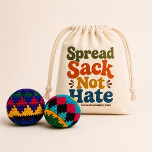 Peut inclure: Deux balles crochet&eacute;es color&eacute;es avec des motifs g&eacute;om&eacute;triques devant un sac &agrave; cordon. Le sac est de couleur cr&egrave;me et porte l'inscription "Spread Sack Not Hate" en plusieurs couleurs. Le site web www.shoplumily.com est imprim&eacute; en dessous.