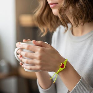 May include: A handmade macrame bracelet featuring a red heart design with yellow wings. The bracelet is made with neon green cord and has adjustable ties. The person is holding a white mug.