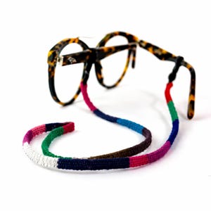 May include: Tortoiseshell eyeglasses with a colorful, woven eyeglass chain. The chain features a pattern of red, pink, green, blue, and white sections. The glasses are set against a white background.