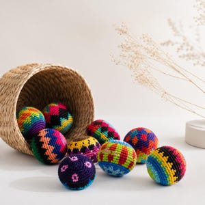 Peut inclure: Plusieurs balles crochet&eacute;es color&eacute;es aux motifs vari&eacute;s, tombant d'un panier en osier. Les balles pr&eacute;sentent des couleurs vives comme l'arc-en-ciel, le rose, le bleu et le jaune. Le panier est en fibres naturelles, sur un fond blanc doux.