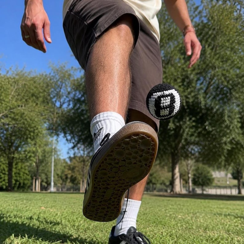 Peut inclure: Un ballon de billard crochet&eacute; noir et blanc est frapp&eacute; en l'air. Le ballon est mis au point, avec le pied et la jambe d'une personne au premier plan. La personne porte des baskets noires, des chaussettes blanches et un short marron. L'arri&egrave;re-plan est un champ herbeux vert et des arbres.