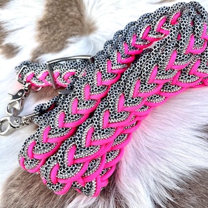 May include: Close-up of a braided dog collar and leash set. The collar and leash feature a black and white pattern with bright pink accents. Silver-colored metal hardware is visible, including a buckle and clasps. The set is displayed on a textured, multi-toned surface.