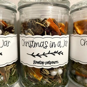 May include: Three clear glass jars filled with dried orange slices, pine needles, cranberries, and white beads. Each jar features a white label with a scalloped edge, black text reading Christmas in a Jar, and the words stovetop potpourri below.