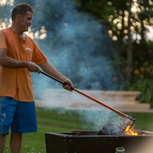 May include: A person uses a long-handled tool to stir a fire in a black metal fire pit. The fire pit is surrounded by green grass and there is smoke rising from the fire.