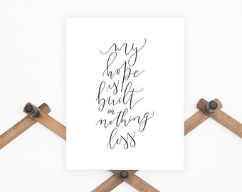 May include: A white framed print with black calligraphy that reads "my hope is built on nothing less". The print is hanging on a wooden coat rack with a blue denim jacket and a gray baseball cap hanging below.