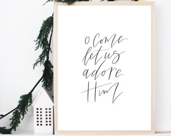May include: A white framed print with black calligraphy that reads "O come let us adore Him".