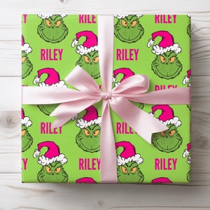 May include: A wrapped gift with green wrapping paper featuring a repeating pattern of a green cartoon character wearing a pink Santa hat. The word "RILEY" is printed in pink. A light pink ribbon is tied around the gift.