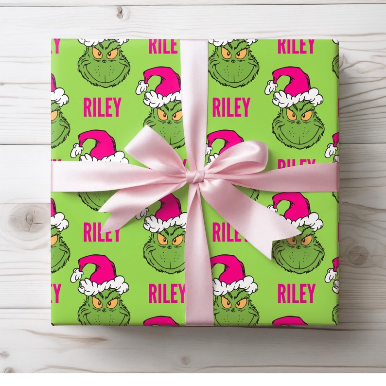 May include: A wrapped gift with green wrapping paper featuring a repeating pattern of a green cartoon character wearing a pink Santa hat. The word "RILEY" is printed in pink. A light pink ribbon is tied around the gift.