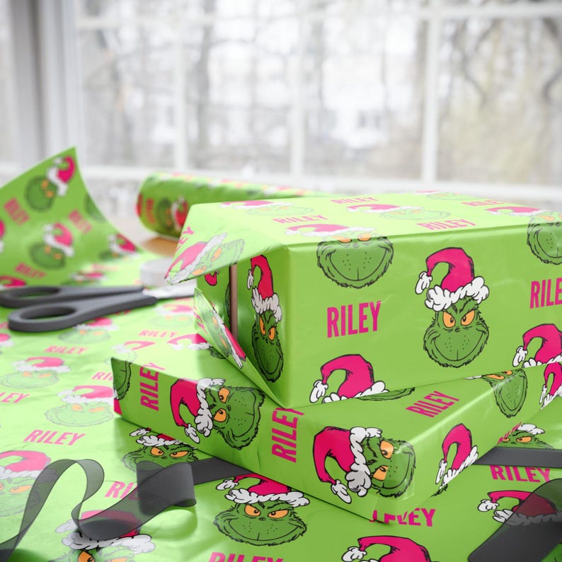 May include: Green wrapping paper with a repeating pattern of a green cartoon character wearing a red and white Santa hat. The name "Riley" is printed on the paper. Several wrapped boxes are visible.