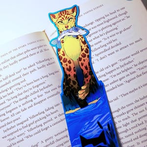 May include: A colorful bookmark featuring a stylized illustration of a cat holding a fish. The cat has yellow fur with brown spots and is set against a blue and green background. The bookmark is placed on an open book.