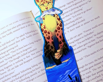 May include: A vibrant bookmark depicting a cat with yellow fur and brown spots, holding a fish. The design is set against a blue and green backdrop. The bookmark is placed on an open book, with text visible.