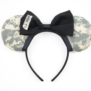 May include: A black headband with two padded ears in a digital camouflage pattern. A black bow is centered on the headband, with a small tag that reads "U.S. ARMY". The ears and bow are the focal point of this novelty item.