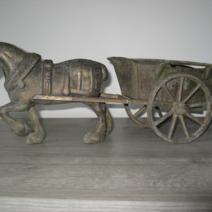 Könnte beinhalten: Ein bronzefarbenes Zierpferd zieht einen Wagen. Das Pferd ist mit Zaumzeug und Geschirr detailliert. Der Wagen hat Speichenräder und ein verwittertes Aussehen, was auf einen antiken oder Vintage-Stil hindeutet. Es ist wahrscheinlich ein Dekorationsartikel.