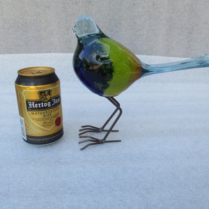 Op de afbeelding: Een blauwe, groene en gele glazen vogelsculptuur met een lange staart en metalen poten. De vogel staat op een wit oppervlak naast een blikje Hertog Jan bier.