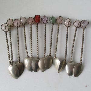 May include: Set of twelve silver teaspoons with twisted handles and heart-shaped bowls. Each handle is topped with a different colored gemstone.