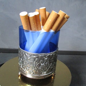 Puede incluir: Un porta cigarrillos de plata con un inserto de vidrio azul. El porta cigarrillos tiene un diseño ornamentado con una escena de personas y edificios. El porta cigarrillos está sobre una superficie dorada.
