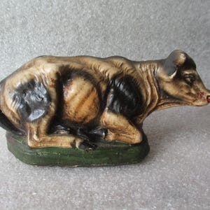 May include: A ceramic figurine of a reclining cow, painted with brown, black, and tan hues. The cow rests on a green base, with a red nose and detailed fur texture. This decorative piece is likely a collectible or a part of a nativity scene.