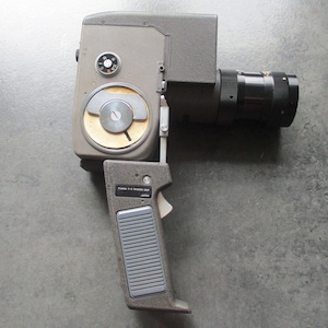 Op de afbeelding: Een vintage Canon 8-14 Trigger Grip 8mm filmcamera met een zwarte lens en een grijs lichaam. De camera heeft een zilveren wijzerplaat met nummers en een zwarte knop. De camera staat op een grijs oppervlak.
