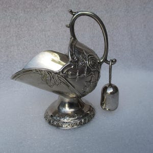 May include: A silver sugar bowl with a handle and a small bell-shaped scoop. The bowl has a decorative floral design and sits on a pedestal base. The handle is arched, and the scoop hangs from a small chain.