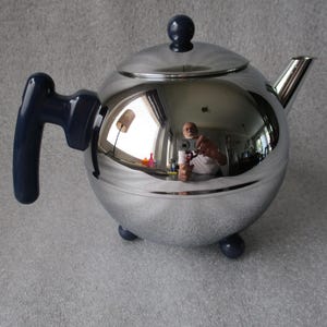 May include: A shiny, spherical teapot with a polished silver finish. It features a dark blue handle, lid knob, and small feet. The teapot's surface reflects the surrounding environment, including a person holding a camera.