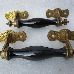 Könnte beinhalten: Zwei antike Schranktürgriffe mit schwarzen Keramikgriffen und verzierten goldenen Metallakzenten.