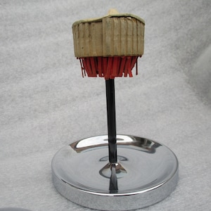 May include: A vintage, chrome-plated, ring holder with a round base and a central post. The post is topped with a circular, paper-covered, ring holder with red fringe.