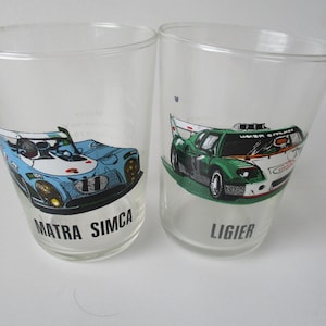 May include: Two clear glass tumblers with racing car designs. The left tumbler features a blue and white Matra Simca car, while the right tumbler features a green and white Ligier car.