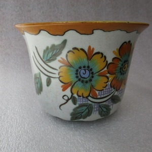 May include: A white ceramic planter with a scalloped orange rim and floral design. The flowers are yellow, orange, and blue with green leaves and stems. The planter has a slightly flared shape.