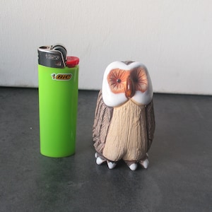 May include: A small ceramic owl figurine with brown and white painted details. The owl has large, round eyes with orange pupils and a white beak. The figurine is standing on a black surface.