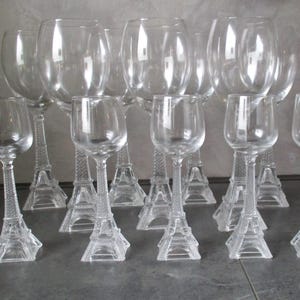 May include: A collection of clear glass wine glasses. The stems are designed as miniature Eiffel Towers. The glasses vary in size, with some being taller and wider than others. The glasses are arranged on a dark surface.