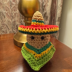 Pode incluir: Uma boneca de crochê usando um sombrero verde, amarelo e vermelho. A boneca tem olhos pretos e um corpo bege.